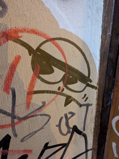 Stylized face drawn with black marker on grey wall, surrounded by other tags.