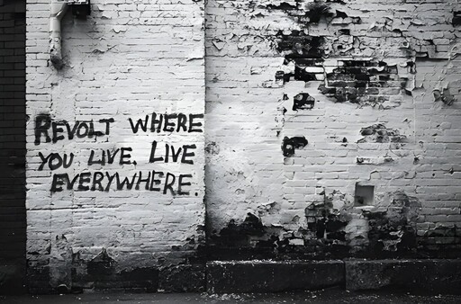 Black-and-white picture of a wall with the graffiti: "Revolt where you live. Live everywhere."